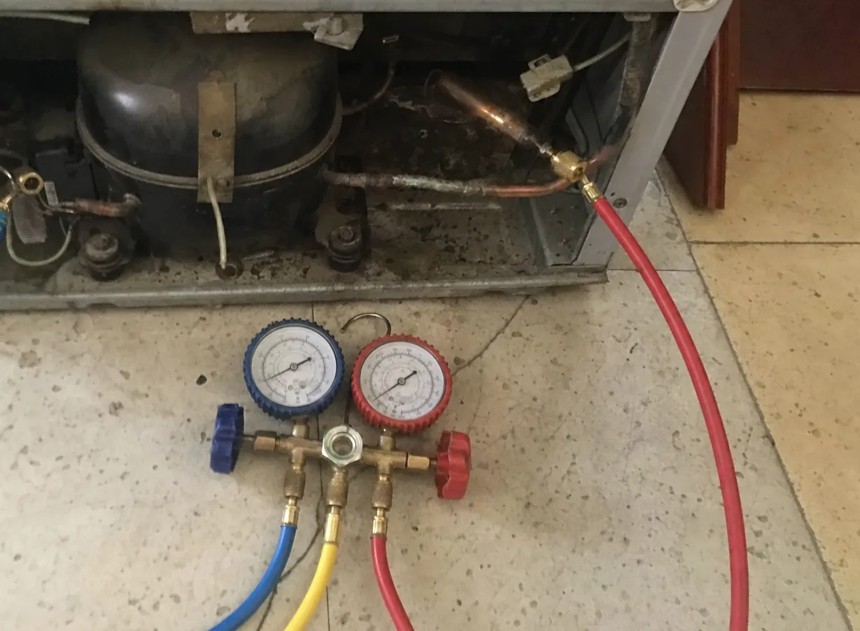 A manifold gauge set on the kitchen floor with a red hose running up into the open compressor compartment of a built-in refrigerator