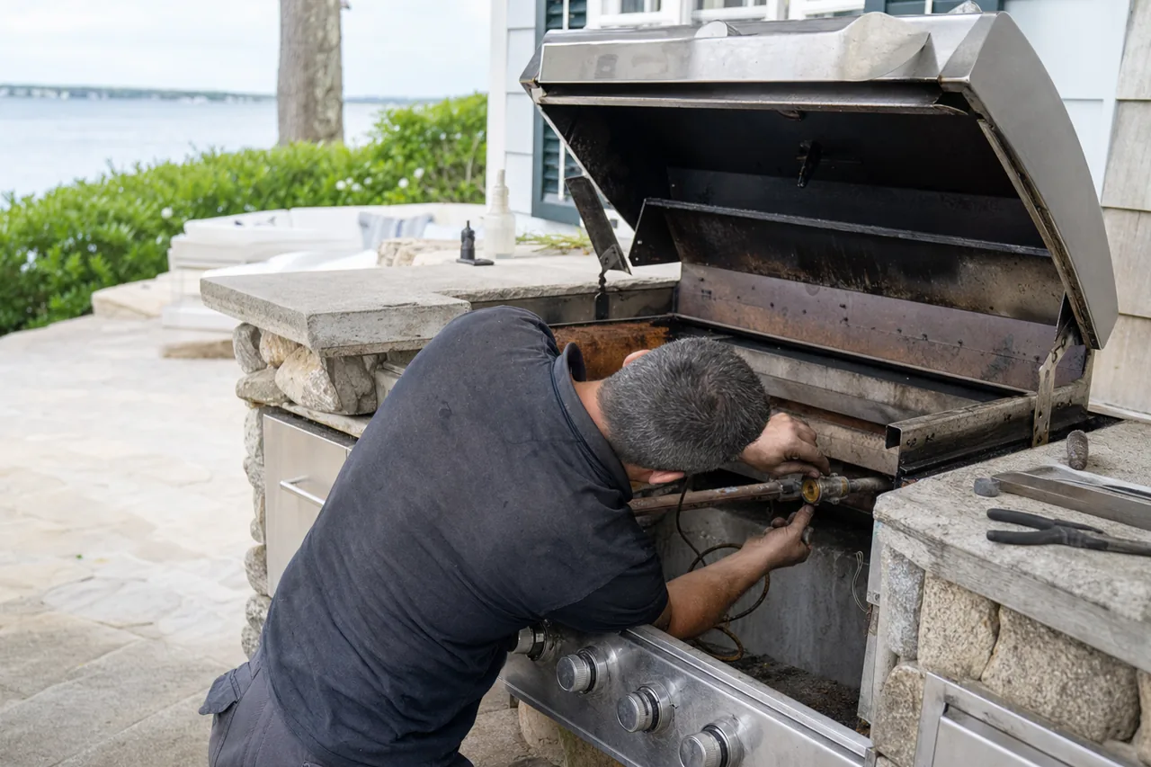 A technician working at the burner and gas connection of a built-in stainless grill, control knobs in the foreground